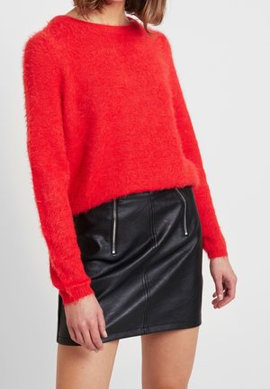 Femme portant un pull rouge duveteux à manches longues associé à une mini-jupe en cuir noir avec deux poches zippées verticales.