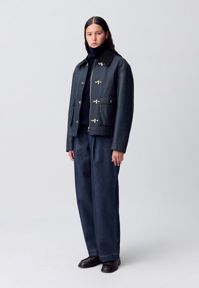 Veste en denim noir avec des attaches dorées, un col noir et des poches avant. Assortie à un pantalon bleu foncé ample et des chaussures noires.