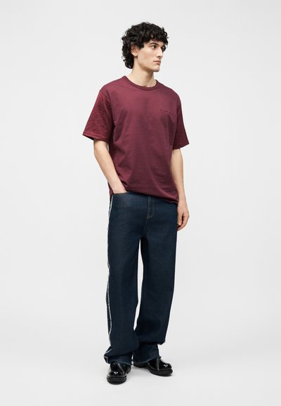 Νέος άνδρας με κυματιστά μαλλιά που φοράει μια φαρδιά μπορντό μπλούζα, σκούρα μπλε τζιν παντελόνια φαρδιάς γραμμής με ρίγες στο πλάι και μαύρα παπούτσια, στέκεται με τα χέρια στις τσέπες.