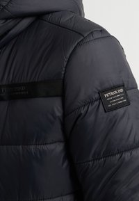 Marineblauwe pufferjack met een gewatteerd ontwerp, een capuchon en een stoffen label op de borst. Glad van textuur met een slanke, lichte uitstraling.
