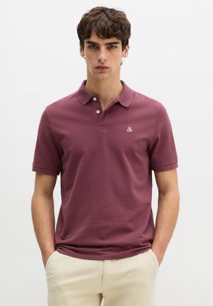 Egy férfi áll zsebre dugott kézzel, bordó rövid ujjú polóingben és bézs színű nadrágban, egyszerű háttér előtt.