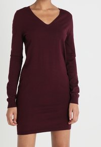 Donna che indossa un vestito maglione attillato, a maniche lunghe, con scollo a V, di un oscuro bordeaux, in piedi contro uno sfondo chiaro e semplice.