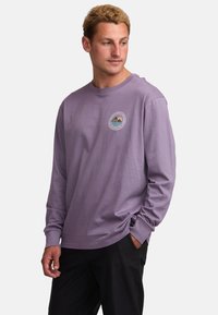 Long-sleeve purple shirt made of soft cotton. Features a round neckline and a circular patch with mountains and water designs.