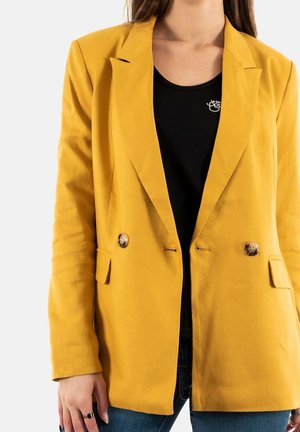 Persoon die een mosterdgele blazer met dubbele knopen draagt over een zwart shirt en blauwe jeans, staand tegen een witte achtergrond.