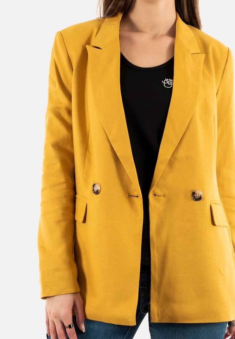 Personne portant un blazer jaune moutarde à double boutonnage sur un haut noir et un jean bleu, debout devant un fond blanc.