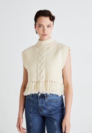 Cream sleeveless turtleneck sweater with a cable knit design, featuring a fringed hem and textured fabric, paired with high-waisted blue jeans.