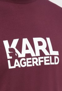 Bordová košile s výrazným bílým logem "KARL LAGERFELD", obsahující siluetu hlavy Karla Lagerfelda, která je integrována do písmene K.