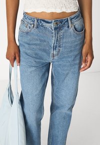 Persona vistiendo jeans de talle alto en azul claro y una blusa corta blanca, sosteniendo una bolsa tote azul claro con una mano contra un fondo sencillo.