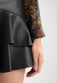 Jupe en cuir noire avec des volants superposés, associée à une manche à motifs colorés. La main est positionnée sur le côté de la jupe.