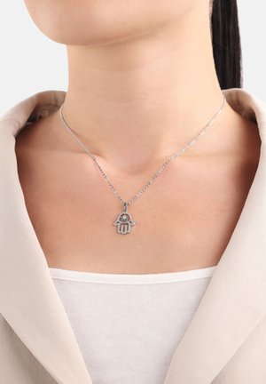 Collana d'argento con pendente a mano di Fatima, adornata con accenti scintillanti, poggiata su una maglietta chiara e sotto un blazer morbido.