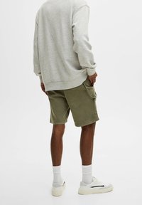 Felpa grigia dal fit rilassato, abbinata a pantaloni corti verde oliva con tasche laterali. Le scarpe da ginnastica bianche e le calze alla caviglia completano l'outfit.
