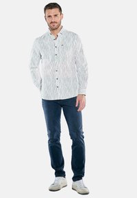 Weiß gestreiftes Langarmhemd mit Button-Down-Kragen, kombiniert mit dunkelblauen Jeans und weißen Turnschuhen. Lässige Passform und Textur sichtbar.