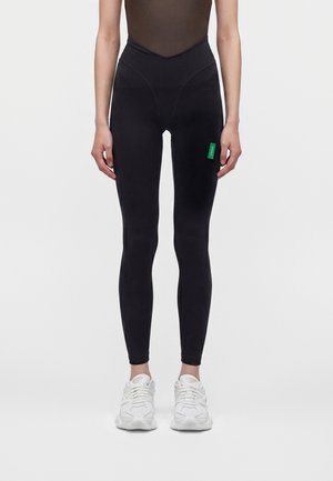 Personne portant un legging noir avec une étiquette verte sur la cuisse et des baskets blanches, debout devant un fond uni.