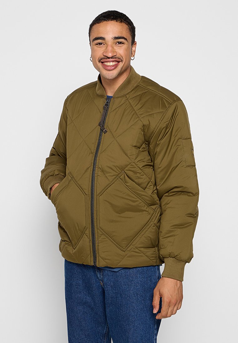 Olivgrüne gesteppte Bomberjacke mit Reißverschluss vorne und gerippten Bündchen. Mit Rautenmuster und einem einzelnen Ärmel-Patch. Trägt man über einem blauen Hemd.