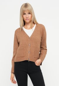 Cardigan marrone in maglia con scollo a V, quattro bottoni metallici, due tasche frontali e polsini a coste. Indossato sopra una camicia bianca.