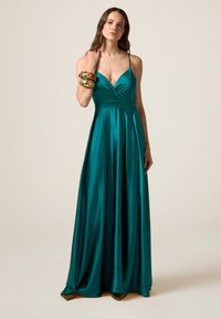 Abito maxi in satin turchese con scollatura a V profonda, corpetto plissettato e gonna fluida. Accentuato da braccialetti colorati al polso.