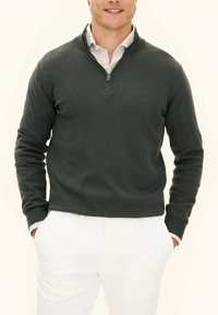 Mörkgrå tröja med dragkedja, ribbade manschetter och en texturerad stickad design, i kombination med ljusa byxor.