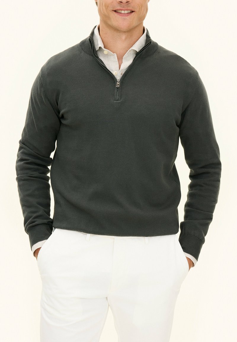 Mörkgrå tröja med dragkedja, ribbade manschetter och en texturerad stickad design, i kombination med ljusa byxor.