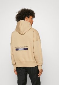 Felpa con cappuccio beige caratterizzata da un design rilassato, polsini a costine e una stampa grafica sulla parte posteriore con il nome del marchio e l'anno di fondazione.