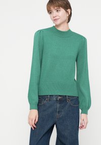 Giovane che indossa un maglione di maglia verde con maniche a sbuffo e jeans di denim blu larghi, in piedi davanti a uno sfondo bianco semplice.