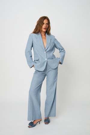 Femme posant en tailleur bleu clair à fines rayures avec pantalon à jambes larges et blazer assorti, portant des talons ouverts bleus avec détail floral.