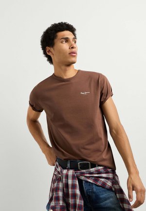 Giovane uomo con una t-shirt marrone e jeans scuri, con una camicia rossa a quadri legata intorno alla vita, che guarda di lato contro uno sfondo semplice.