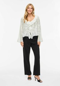 Lahka, polprosojna bluza z naborki pri ovratniku in razširjenimi rokavi, kombinirana s črnimi ravnimi hlačami in črnimi sandalami z visokim peto.