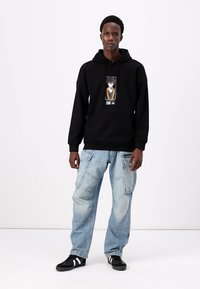Svart hettegenser med grafisk trykk av en karakter foran, kombinert med løse lyseblå cargo jeans og svarte joggesko.