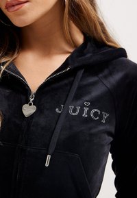 Juodasis velūrinis užtrauktukas su deimantiniu akcentu, rašantis "JUICY", ir širdelės formos užtrauktuko laikiklis. Minkšta tekstūra, gobtuvas.
