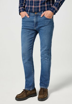Niebieskie dżinsy w slim fit, z standardowym stanem, pięcioma kieszeniami i kontrastowymi szwami. W połączeniu z brązowymi zamszowymi butami.