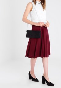 Czarna torebka w stylu kopertówki z łańcuszkiem, wykonana z gładkiego materiału; noszona z białą koronkową bluzką i bordową spódnicą plisowaną. Czarne szpilki.