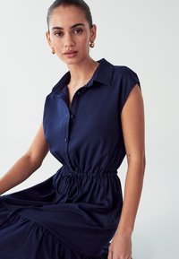 Tamsiai mėlynas suknelė trumpomis rankovėmis, su mygtukų užsegimu priekyje, elastinga liemens linija ir raukšlėtu apvadu. Pagaminta iš lygios medžiagos su švelniu tekstūru.