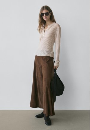 Femme portant un haut beige texturé à manches longues, une jupe large marron, des bottes noires et des lunettes de soleil, tenant un sac à main noir sur un fond uni.