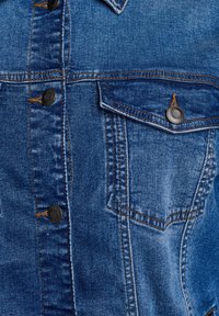 Traper jakna s tamnoplavom tkaninom, dugmadi na zatvaranju i jednim džepom na grudi. Vidljivo šivanje i teksturirana površina pojačavaju dizajn.