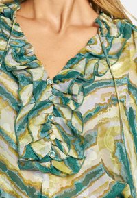 Blusa fluida in verde e giallo con colletto a volant, tessuto strutturato, dettagli con bottoni e un motivo ondulato che crea un aspetto stratificato.