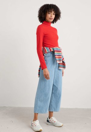 Rode geribbelde coltrui, lichtblauwe wijde jeans en witte sneakers met kleurige accenten, aangevuld met een multicolor gebreide sjaal die om de taille is geknoopt.