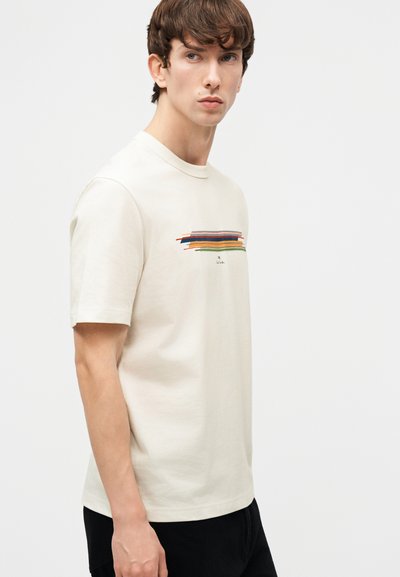 Νεαρός με κοντά καστανά μαλλιά στέκεται πλευρικά, φορώντας λευκό μπλουζάκι με πολύχρωμο οριζόντιο σχέδιο από πινελιές στο στήθος.