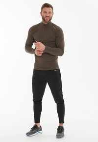 Lange mouwen, ritsbovenkant in donkerbruin, gecombineerd met zwarte, taps toelopende joggers. Bevat vochtregulerende stof en reflecterende accenten op de kleding.