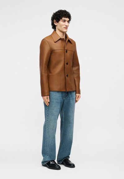 Uomo in piedi che indossa una giacca di pelle marrone con bottoni, jeans blu e scarpe di pelle nere su uno sfondo chiaro e semplice.