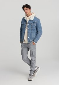 Giubbotto di denim con colletto in pelliccia sintetica, felpa beige sotto, e pantaloni cargo grigi. Scarpe da ginnastica grigie completano l'outfit.