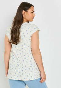Blusa ligera de color crema con estampado floral y mangas cortas, que presenta pequeñas flores azules y detalles verdes. La parte posterior tiene una costura sutil y un dobladillo redondeado.