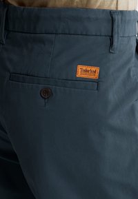 Tummansinisen väriset chinot, joissa on sileä pinta, ruskealla nahkaisella Timberland-tunnuksella, yksittäinen nappisulku ja sivutasku.