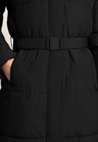 Geacă puf neagră cu talie peptită, având o textură netedă, un design matlasat și buzunare laterale. Stil minimalist cu accesorii ascunse.