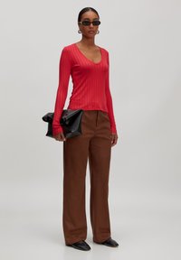 Rotes geripptes Langarmshirt mit V-Ausschnitt, kombiniert mit braunen weiten Hosen. Das Model hält eine schwarze gefaltete Clutch. Schwarze Sonnenbrille getragen.