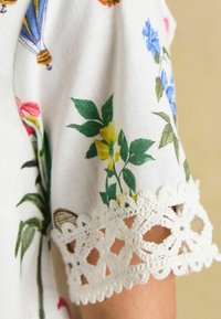 Floral print blouse featuring a white background, colorful flowers, and intricate lace trim on the sleeves, showcasing a detailed design.