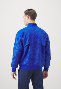 Blauwe sportjack met een textuur ontwerp, ribgebreide manchetten en zoom, en een hoge kraag, gecombineerd met donkere broek. Het patroon bevat geometri sche accenten.