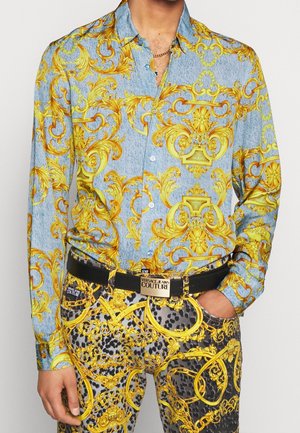 Homme portant un pantalon et une chemise bleu et noir avec des motifs baroques dorés élaborés et une ceinture noire avec une boucle dorée « Versace Jeans Couture ».