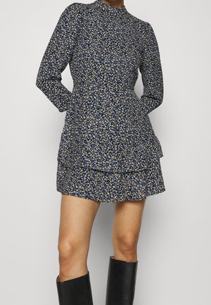 Donna che indossa un vestito nero corto a maniche lunghe e collo alto, con motivo floreale bianco e blu e stivali neri al ginocchio.
