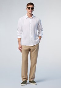 Camicia bianca in lino con bottoni, pantaloni beige in lino e sneakers verdi. Il modello indossa occhiali da sole scuri. Design semplice con una vestibilità rilassata.