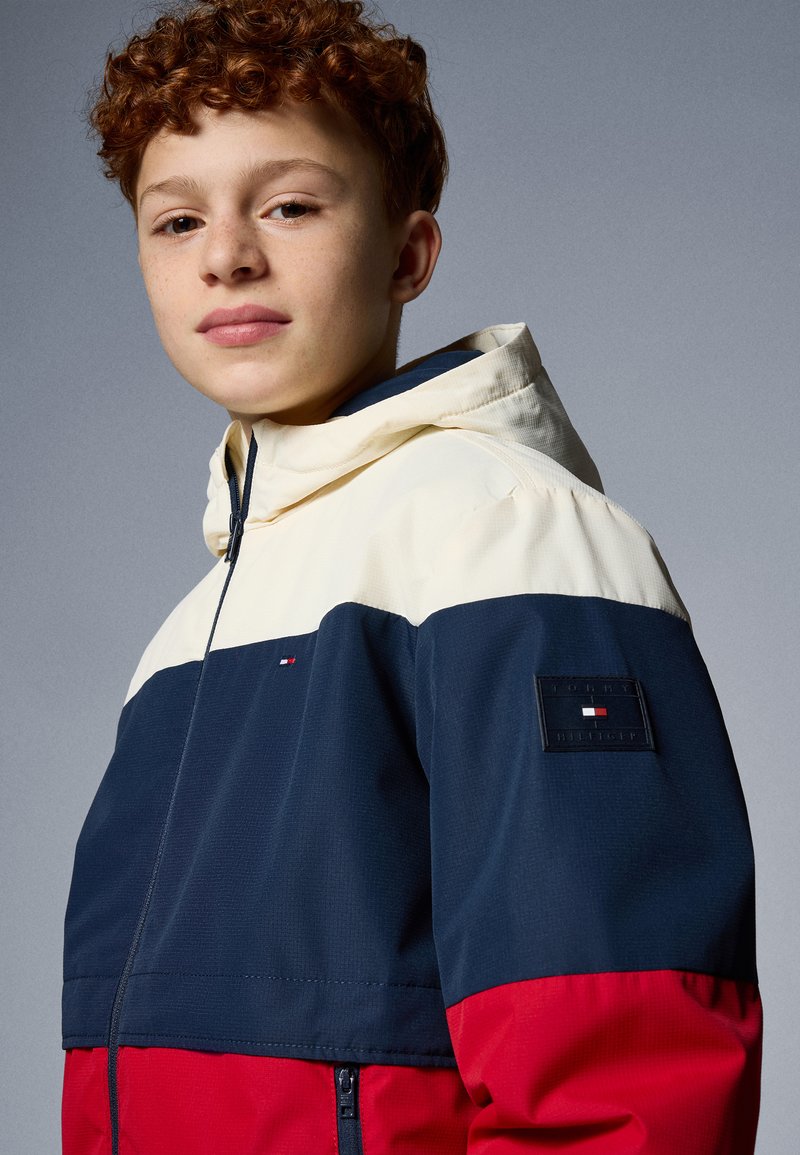 Kleurgeblokte windbreaker in marineblauw, crème en rood. Heeft een ritsluiting, capuchon en textuurstof. Inclusief een logo patch op de mouw.